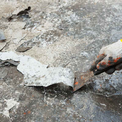 Gloved hand scrapes peeling coating from concrete slab using putty knife during surface preparation work
