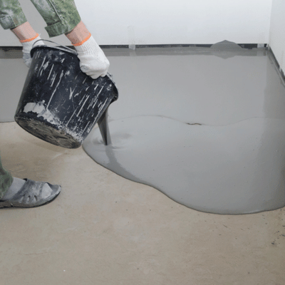 Worker pours self-leveling compound from bucket onto concrete slab, forming even base before epoxy finishing