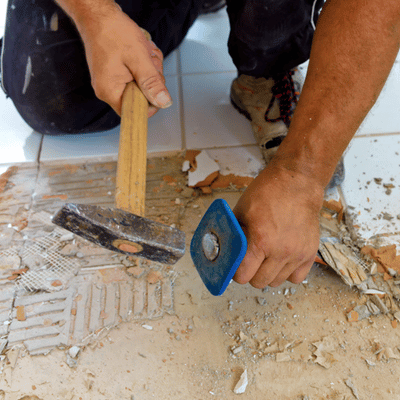 Hands use hammer and scraper to remove tile mortar, continuing concrete stripping on floor area